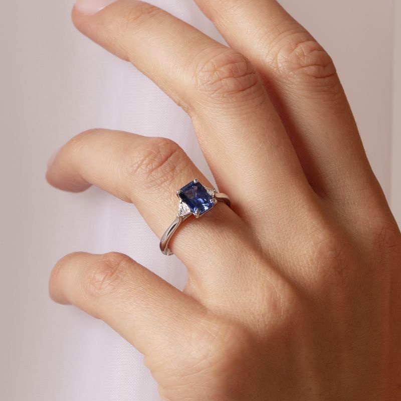 Platinum three stone engagement ring worn on a model's hand, featuring an emerald cut blue sapphire with trilliant cut diamond side stones in claw settings.